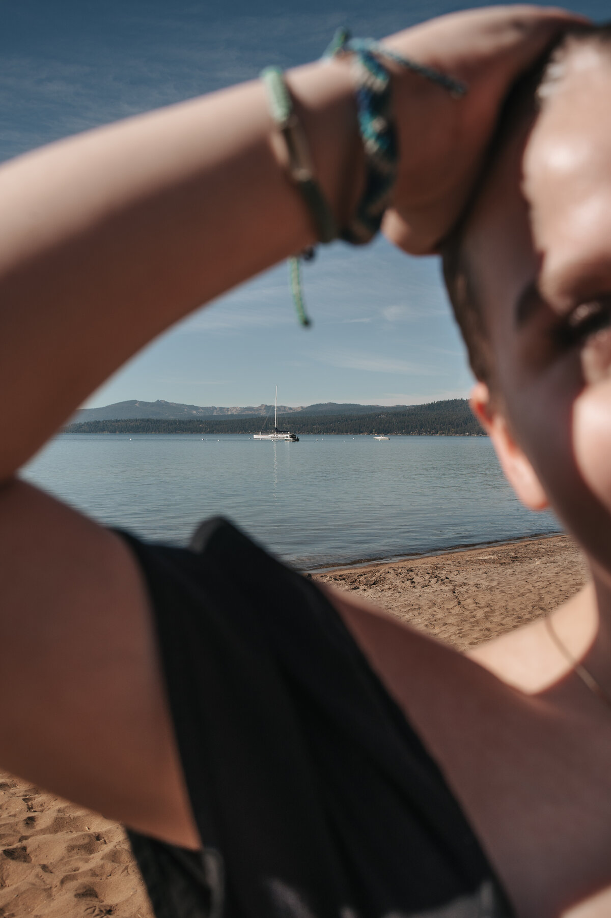Person wearing turquoise bracelets gazing at a sailboat on a calm lake from a sandy Lake Tahoe beach on a sunny day, captured in a serene and scenic outdoor lifestyle photography session.