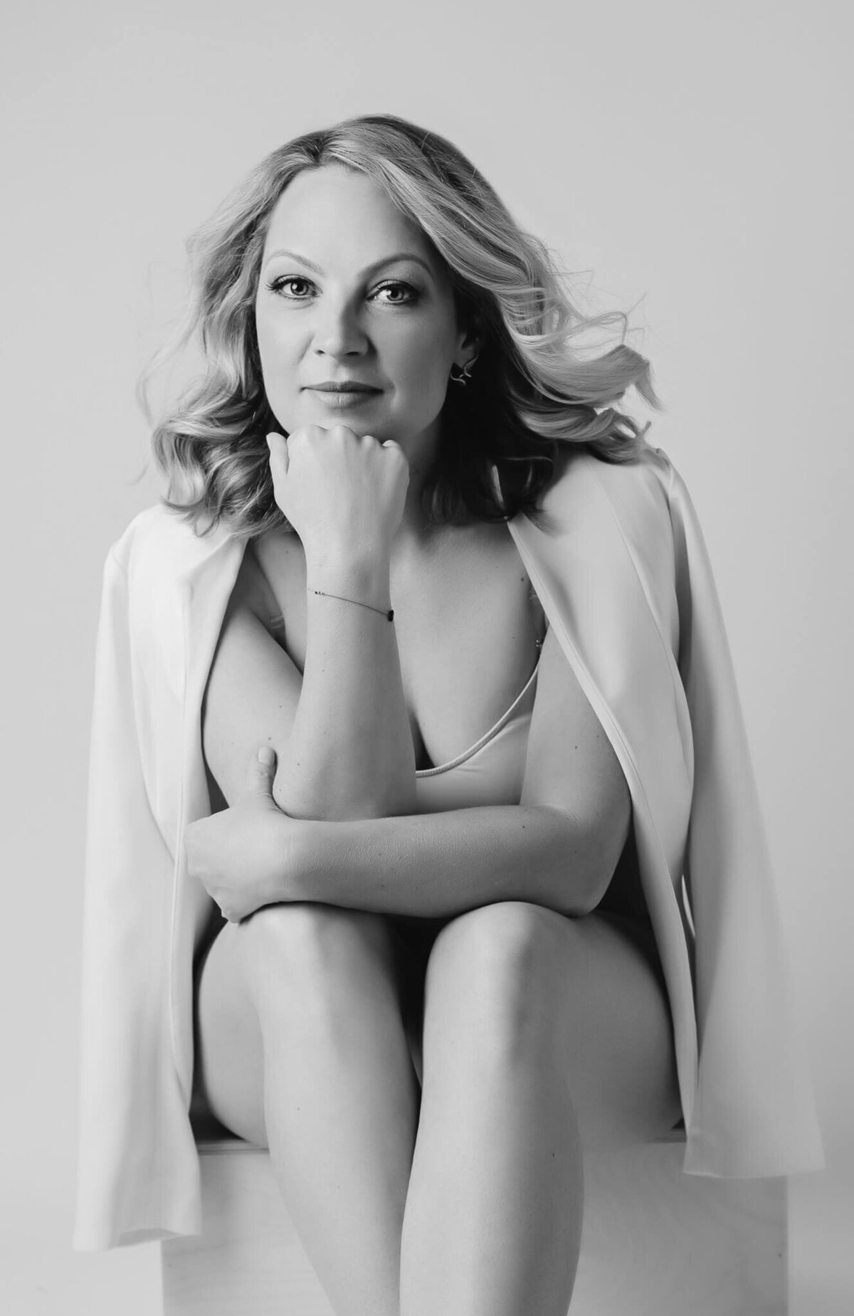 Black and white portrait where you can see a beautiful blonde woman in her 40's posing in an elegant way, she is wearing a coat and a bodysuit and is sitting on a chair