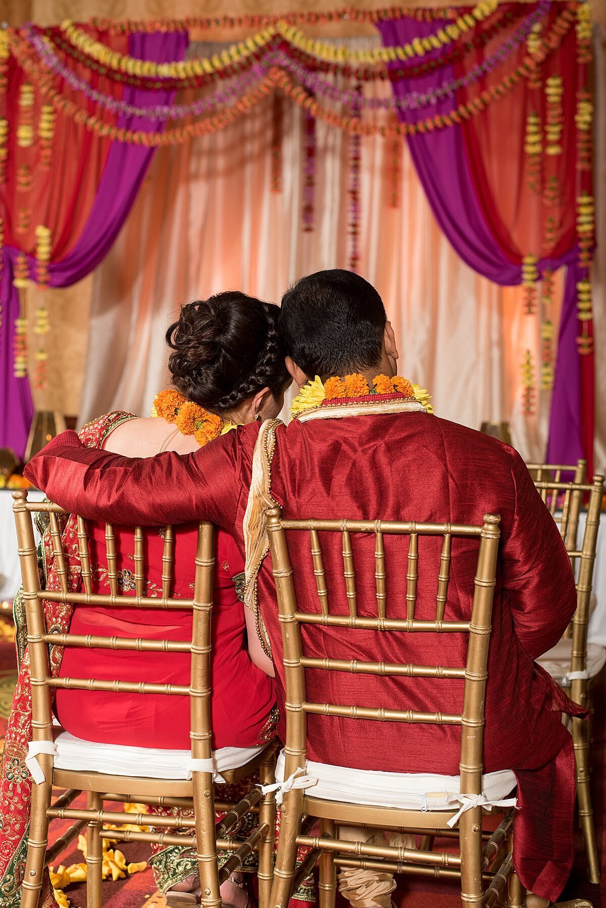 Indian Bride and Hindu Groom sitting in gold chiavari chairs looking at their magenta and red mandap decorated with yellow, purple and orange floral garlands in Nashville, TN