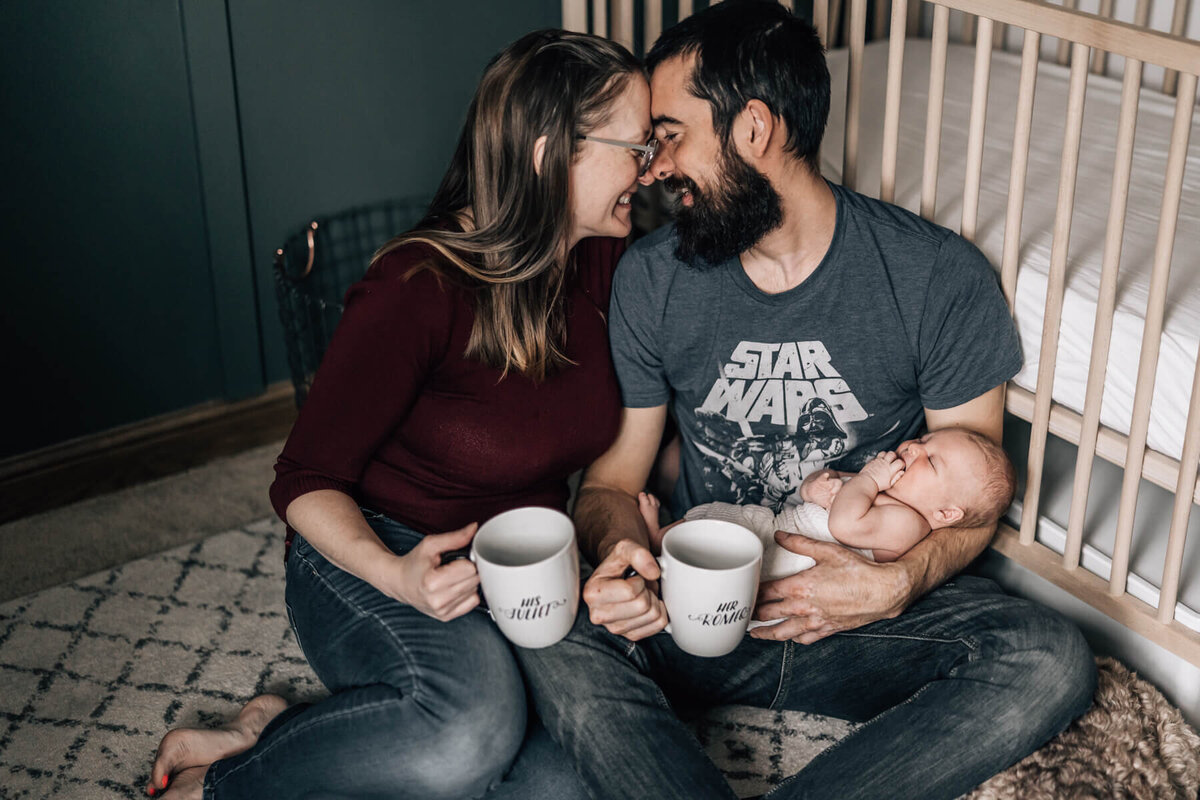 A couple cradling their baby while sitting on the floor of their nursery, holds Romeo and Juliette coffee mugs and looks into each other's eyes.