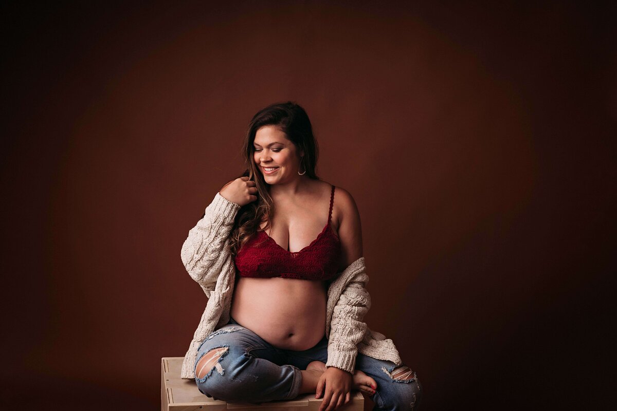 Mom in a casual, hipster outfit featuring blue jeans, a red bra, and a cardingan falling off her shoulders adding a comfortable touch. She looks relaxed  and care-free in this studio maternity photo by Mobile, AL Maternity Photographer. The mom is sitting on a box, with her left foot resting on the box, as she plays with her hair and gives a gentle smile.