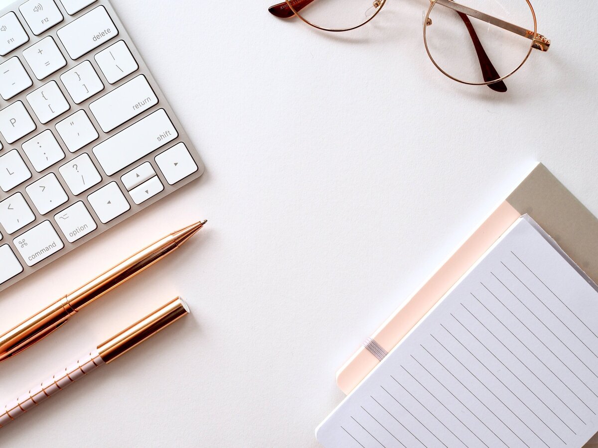 A styled flatlay with a white keyboard, gold pens, a notebook, notepad, and glasses.