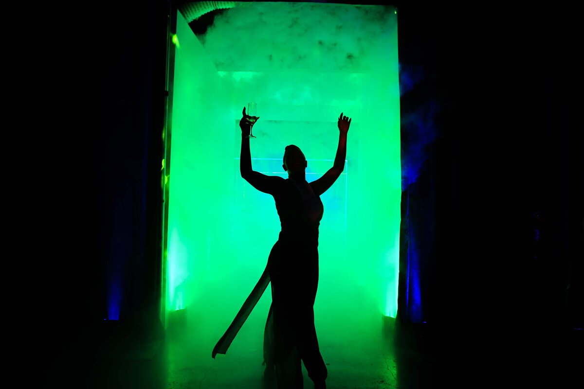 A women holding a drink is silhouetted against green fog in doorway.