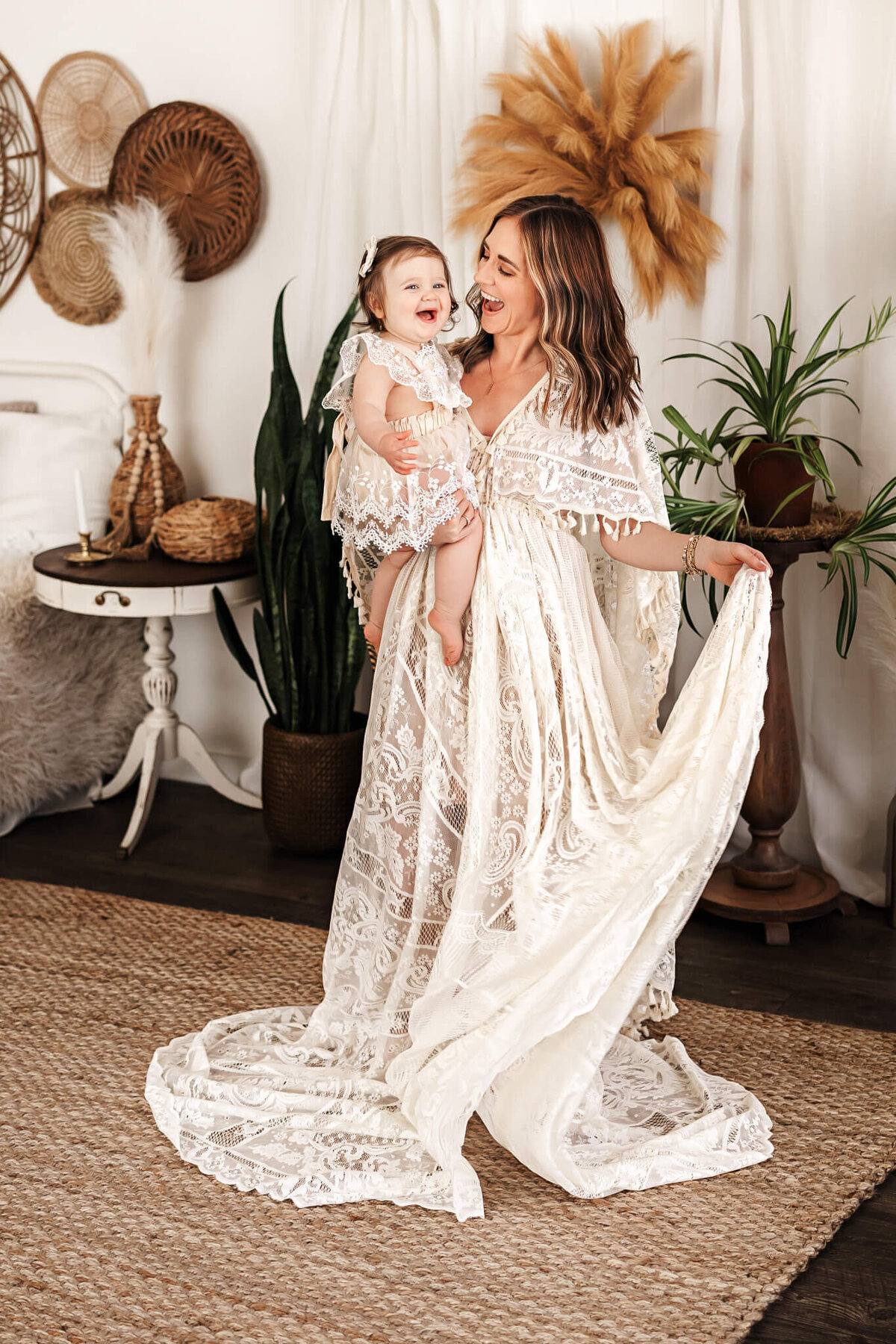 In a warmly bohemian room adorned with plants and woven decor, a portrait photographer captures the essence of family photography. A woman in a flowing lace gown holds her smiling baby, both draped in lace, immortalizing this precious moment amidst the elegance of Bloomington, Illinois.