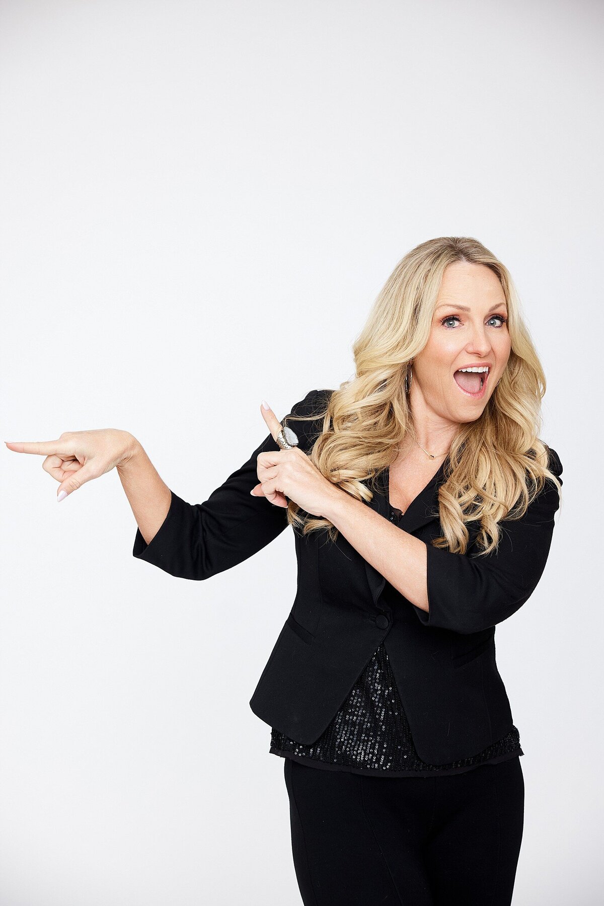 Fun and professional studio headshot of a woman with long blonde curls in a black blazer, smiling and pointing with enthusiasm.