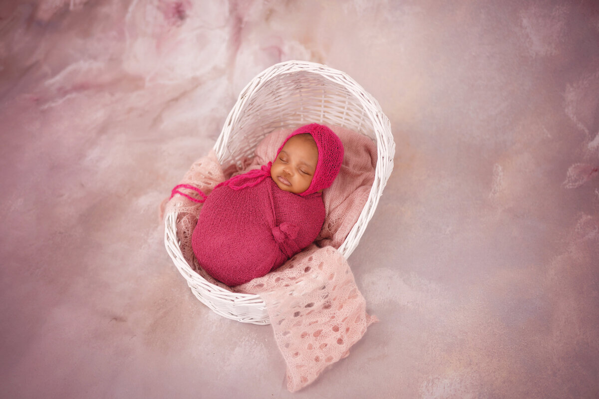African American baby girl in a white basket on a pink floral background.  Baby is wrapped in magenta and also is wearing a magenta bonnet.  She is asleep.