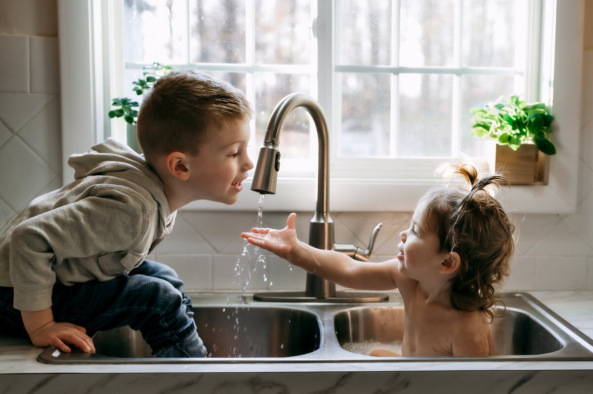 Two young brothers playing in the kitchen sink during an in-home photo session, warm natural light coming through the window, candid and cozy family moment.