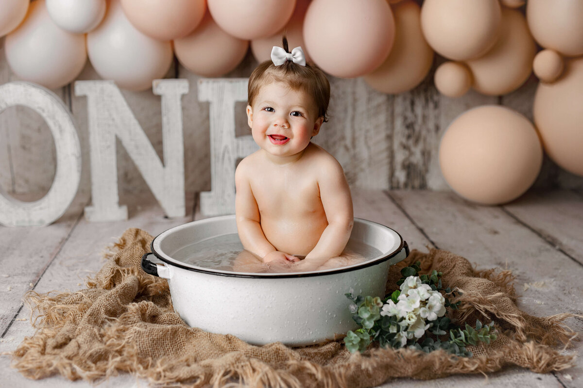 11 month old girl taking a bubble bath with balloons and pink balloons behind her during her one year old session