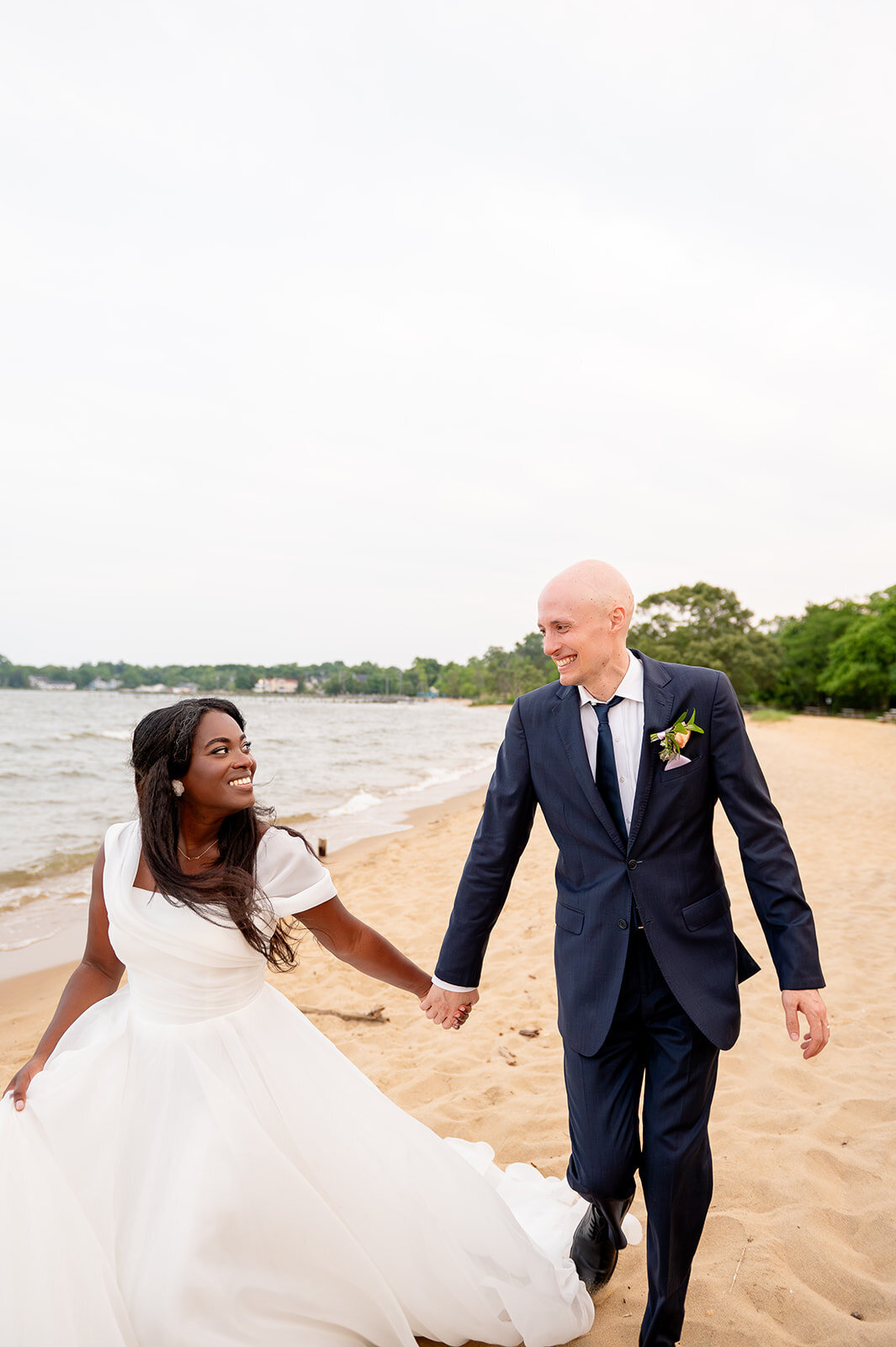 Bride and groom walking hand in hand along a sandy beach, with the bride in a flowing gown and the groom in a navy suit. At the Chesapeake  Bay Foundation.