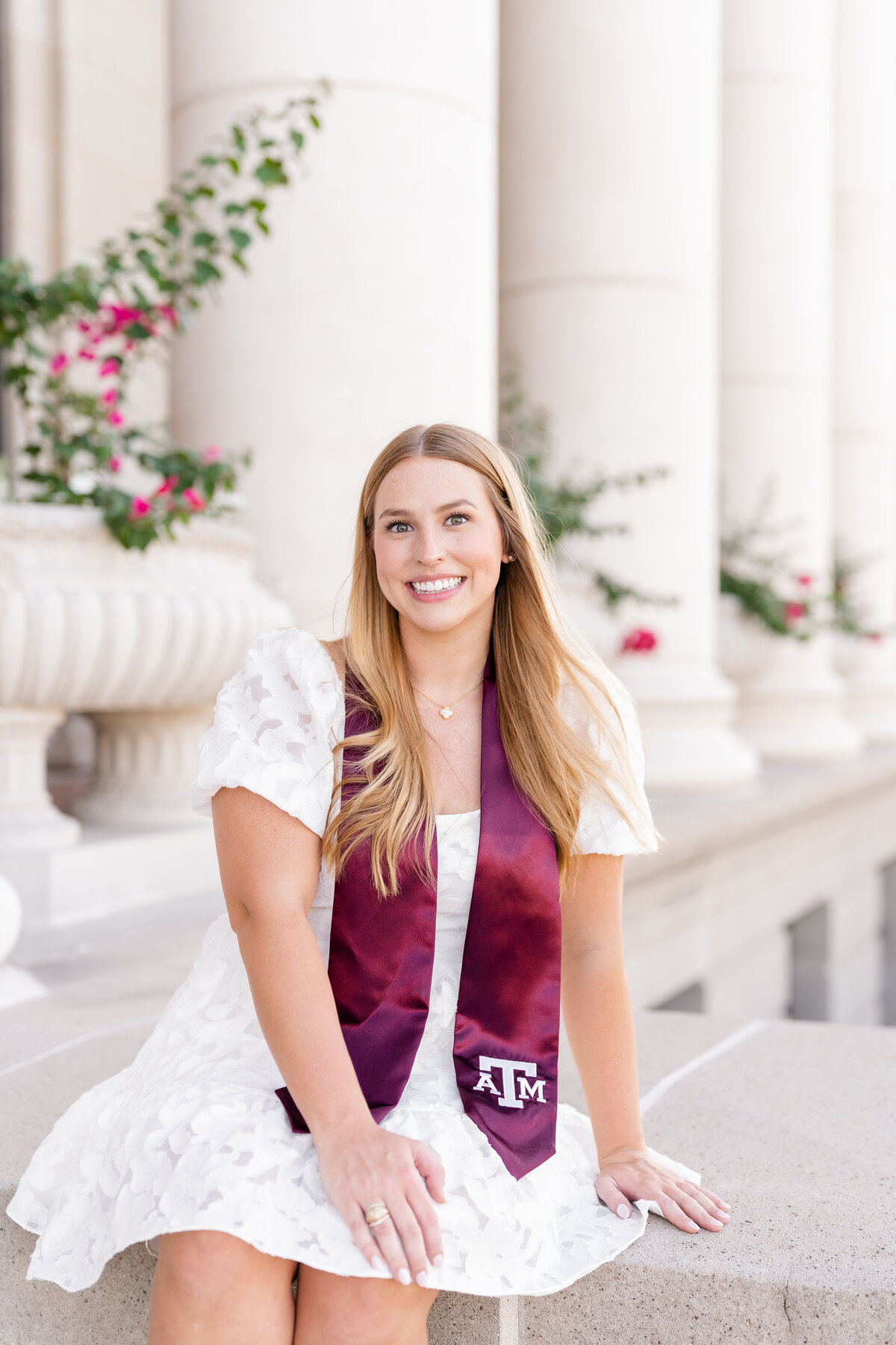 Texas A&M senior girl sitting in front of Administration Building wearing white dress and Aggie stole and smiling