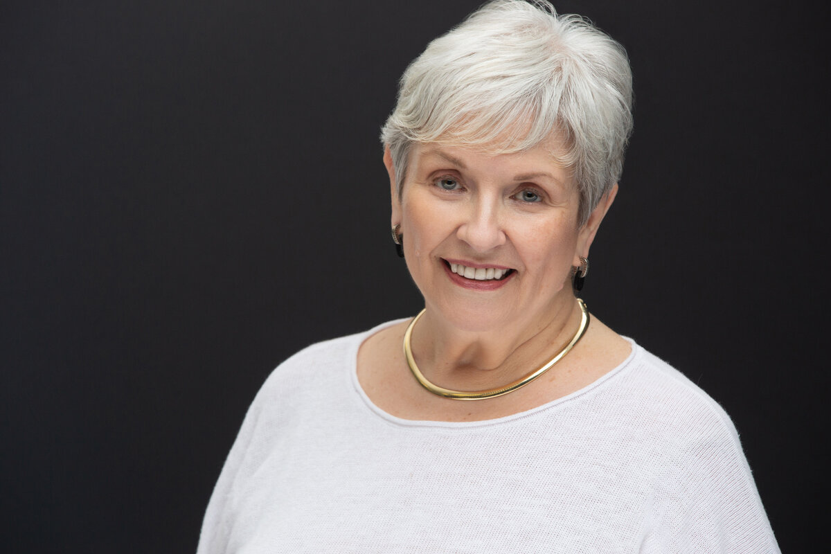 A mature white woman with white hair poses for a headshot on a dark black background at Janel Lee Photography studios in Cincinnati Ohio