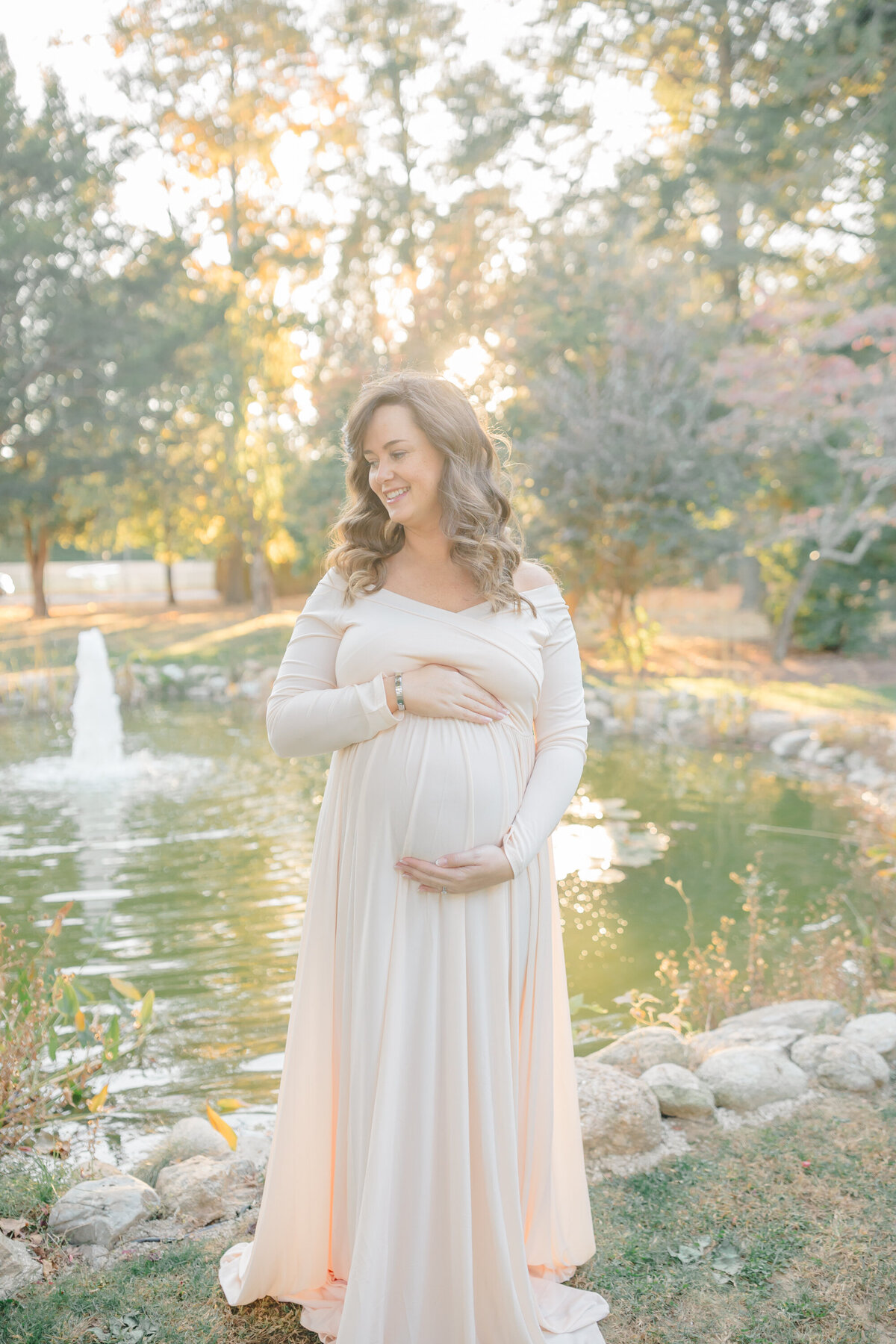 a soft image of a mother to be holding her pregnant belly and standing near a pond, the sun comes behind her hair lighting her up in a gorgeous golden light