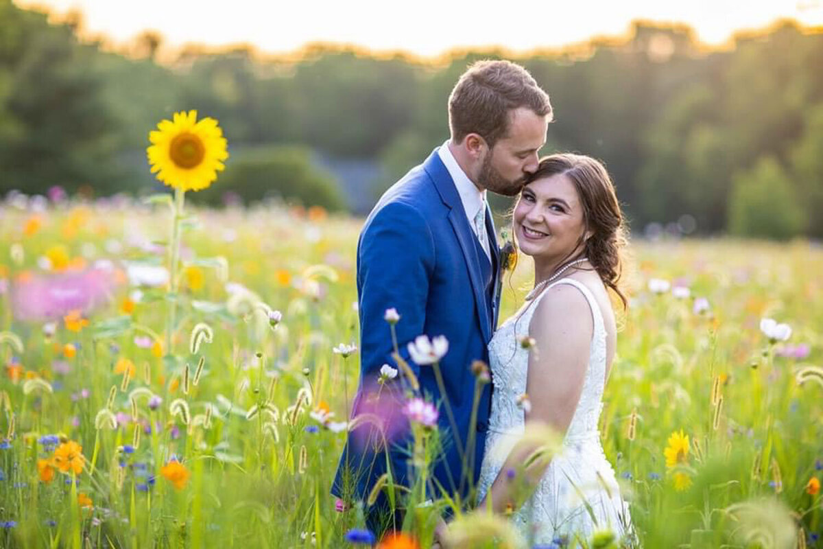 Groom kissing bride’s forehead in a field of sunflowers and wildflowers