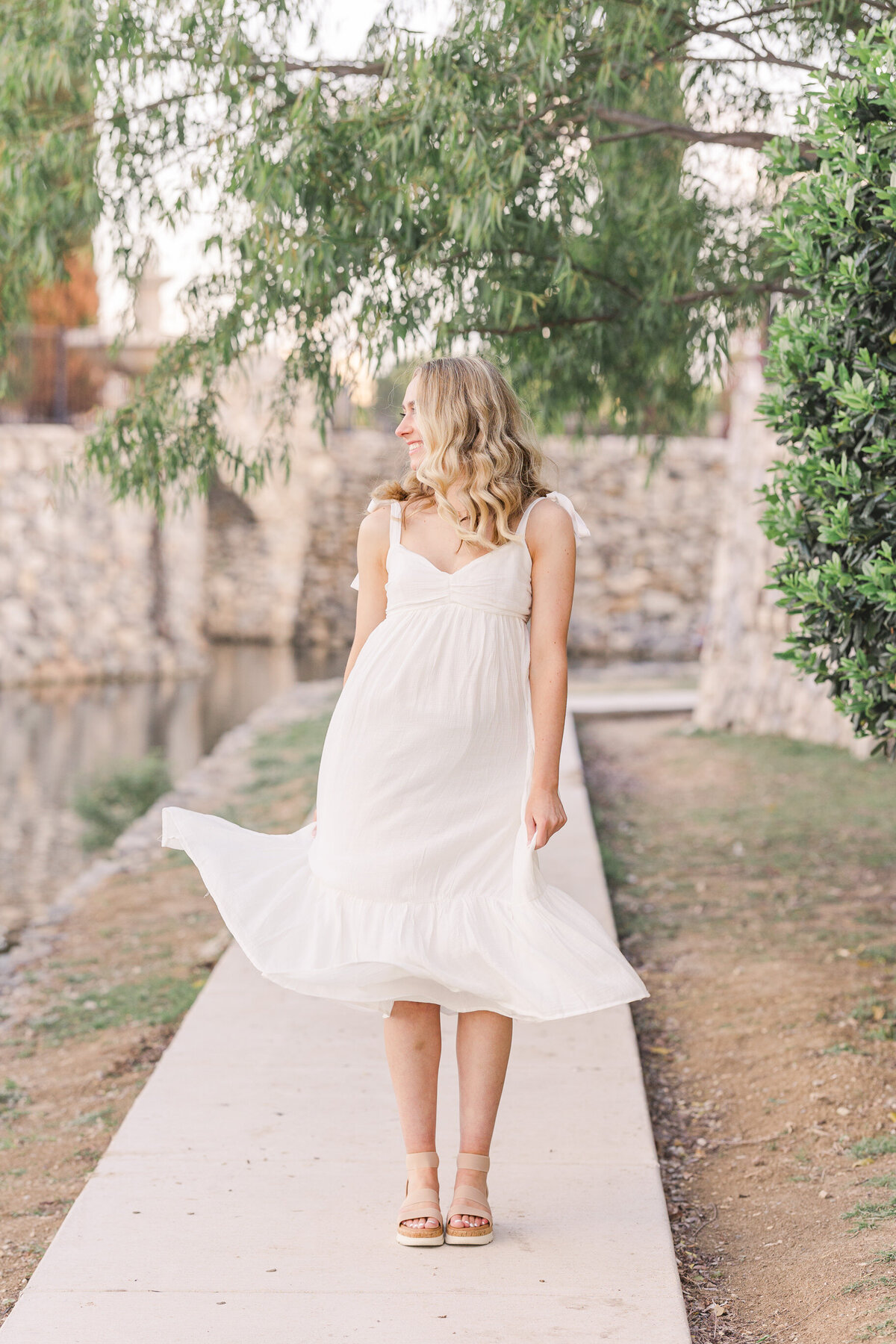 A light and airy senior portrait of a high school graduate twirling in a white dress under the trees at Adriatica Village. Taken in McKinney, TX by Jennifer Lynn Photography, a Dallas Fort Worth senior photographer specializing in timeless and joyful portraits.