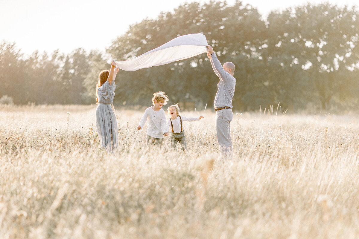 A family photo of mom an dad holding a blanket up in the air as their boys run under in the middle of a DFW field.