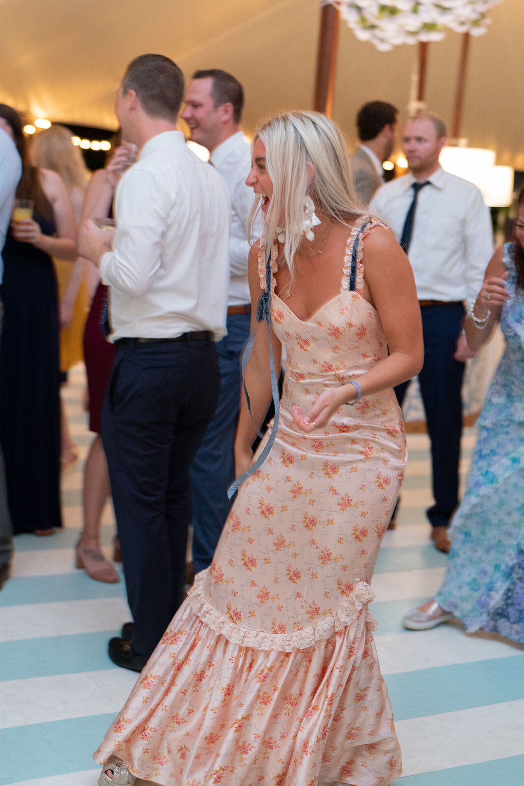 As the vibrant scene unfolds, the setting of this lively gathering at The Hamptons, New York, captures the essence of luxury and elegance. Photographer Bonnie Sen beautifully captures the energy of the moment as a blonde woman in a floral dress takes center stage with her energetic dance moves. To complement the photography, videographers East West Films also skillfully capture the enchanting ambiance of the event. The elegantly dressed individuals in semi-formal attire, immersed in the jubilant spirit, are surrounded by conversations flowing and glasses clinking in the well-lit and lively venue, enhancing the festive atmosphere even further.