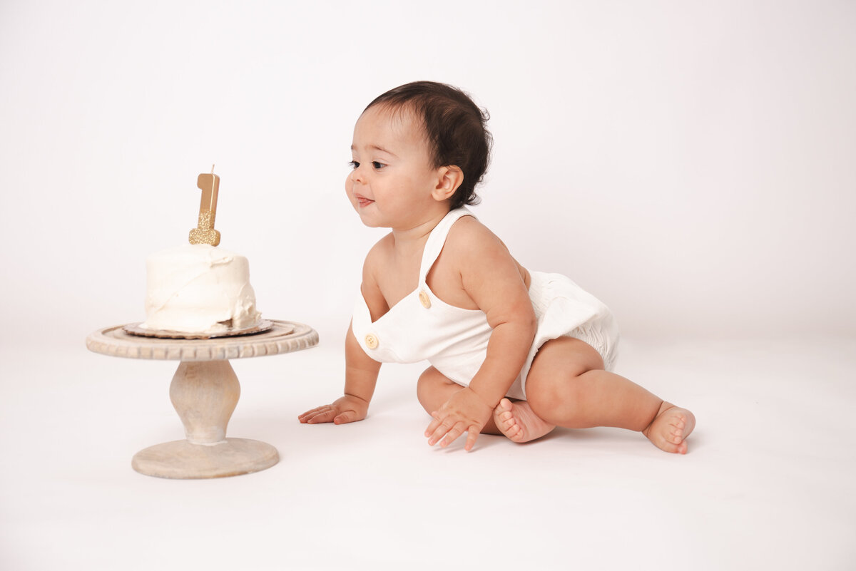 A one-year-old baby in a white romper curiously looks at a small birthday cake with a gold ‘1’ candle during a minimalist cake smash photography session in Connecticut. Captured by Nicole Mele Photography.