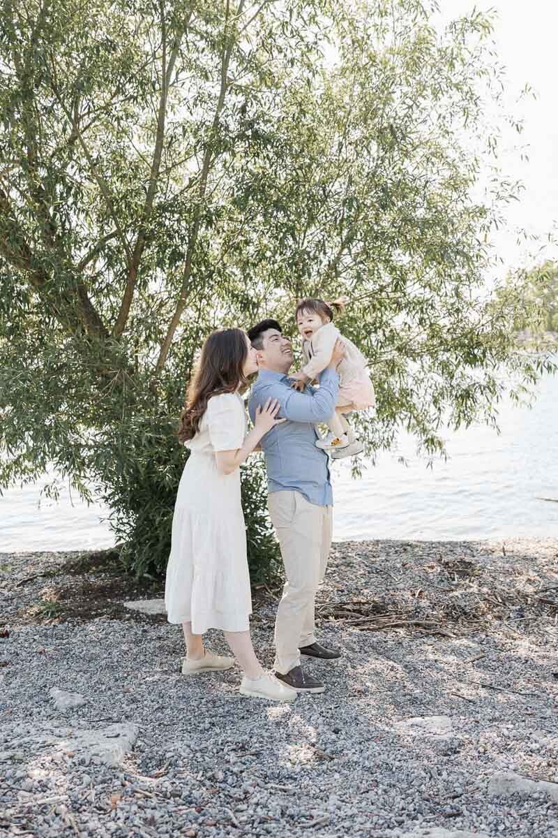 A family of three near a tree by the water; the father holds a young child while the mother stands beside them on a rocky shore—a perfect shot for a Toronto family photographer capturing summer beach photos.