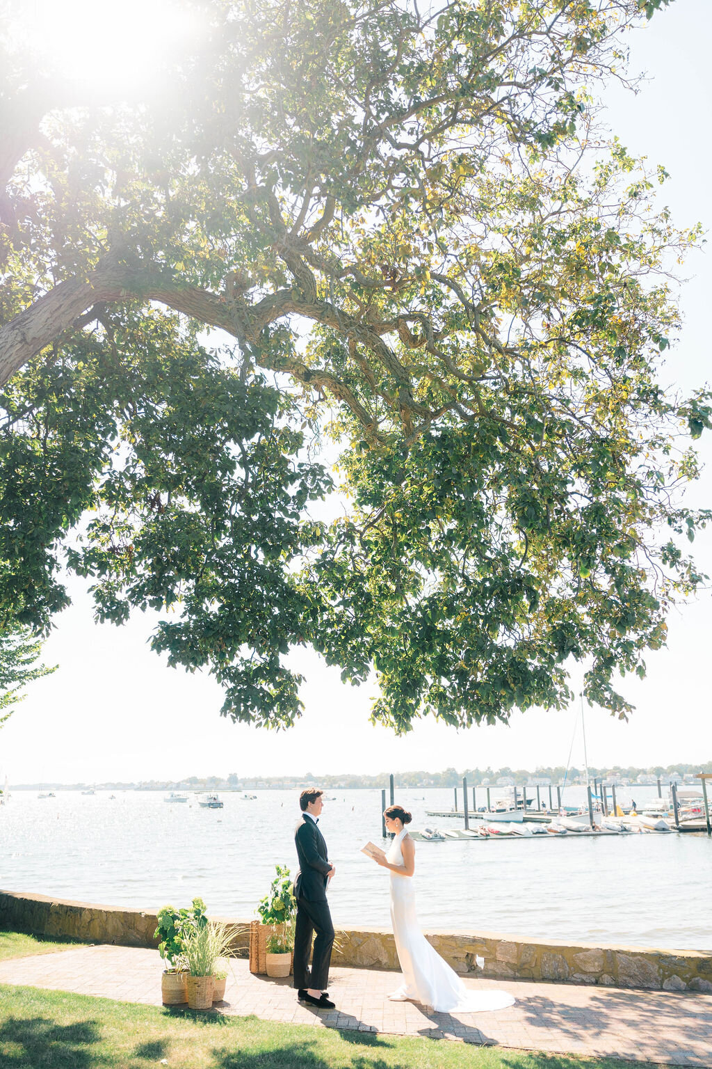 Rhode Island Elopement | A couple stands under a large, green tree by a serene lakeside, exchanging vows. Sunlight filters through the leaves.