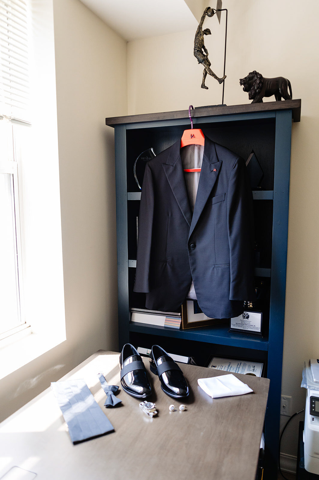 A groom's wedding attire displayed in a room with natural light; a navy blue suit hanging on a shelf with a white dress shirt, shiny black dress shoes, a tie, cufflinks, and a pocket square neatly arranged on a desk