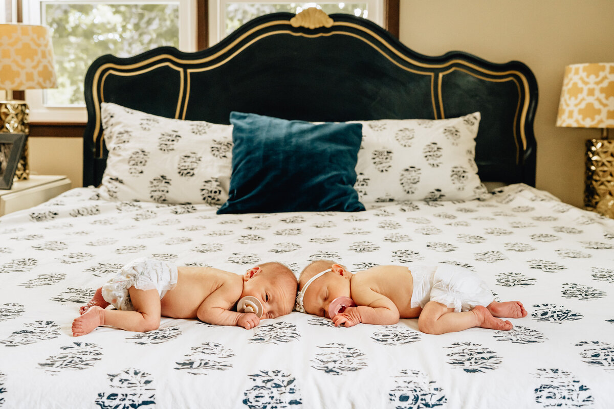Two newborn twins rest peacefully on a large bed, each wearing a diaper and a pacifier.