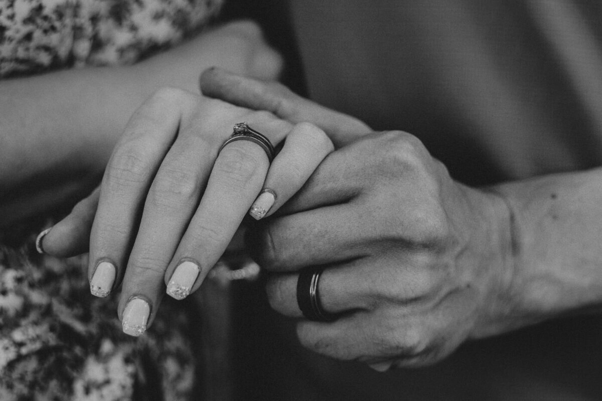Black-and-white detailed photograph of intertwined hands showcasing an engagement ring and wedding bands.