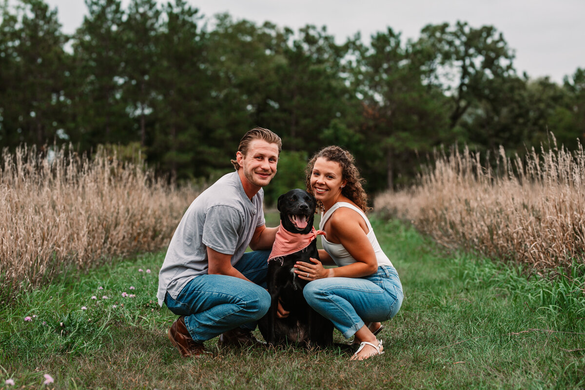 man woman and black lab smile at camera in a field