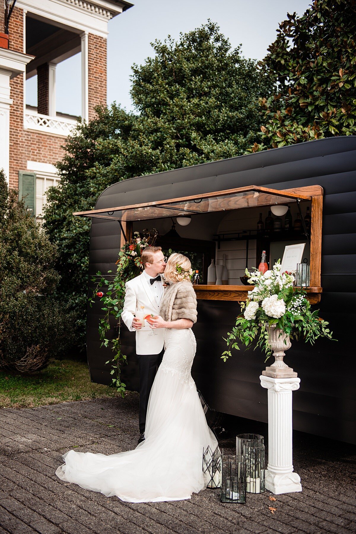 A groom in a tuxedo with a white jacket kisses the bride in front of a black bar trailer. The bride has a gown with a long train with a fur wrap. They are holding signature cocktails next to a white pillar holding a stone planter with a large floral arrangemement with white flowers  and cascading greenery at Rippa Villa.