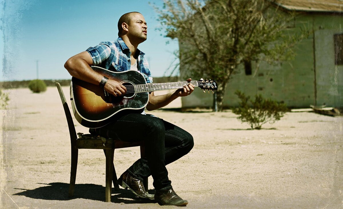Male musician portrait Tebey wearing plaid shirt sitting against wood chair playing guitar desert buildings behind