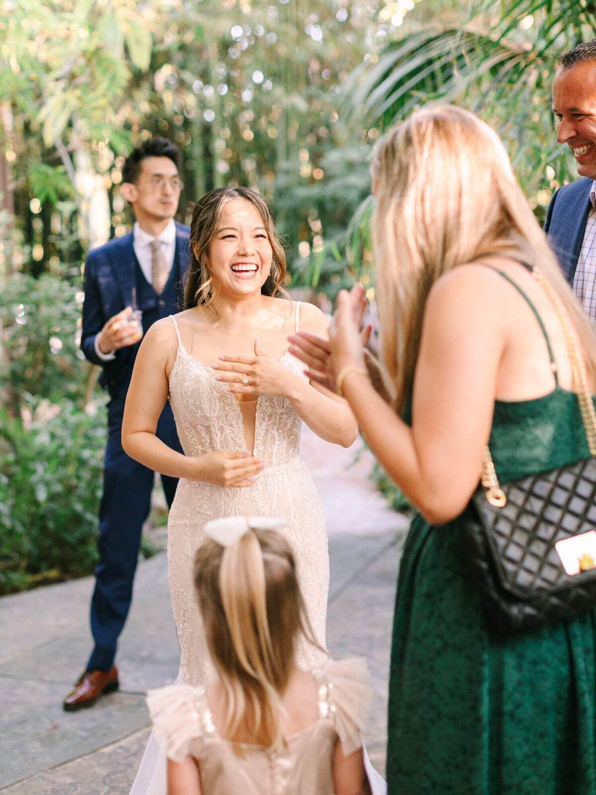A bride in a lace wedding dress laughs joyfully surrounded by guests in a lush garden.
