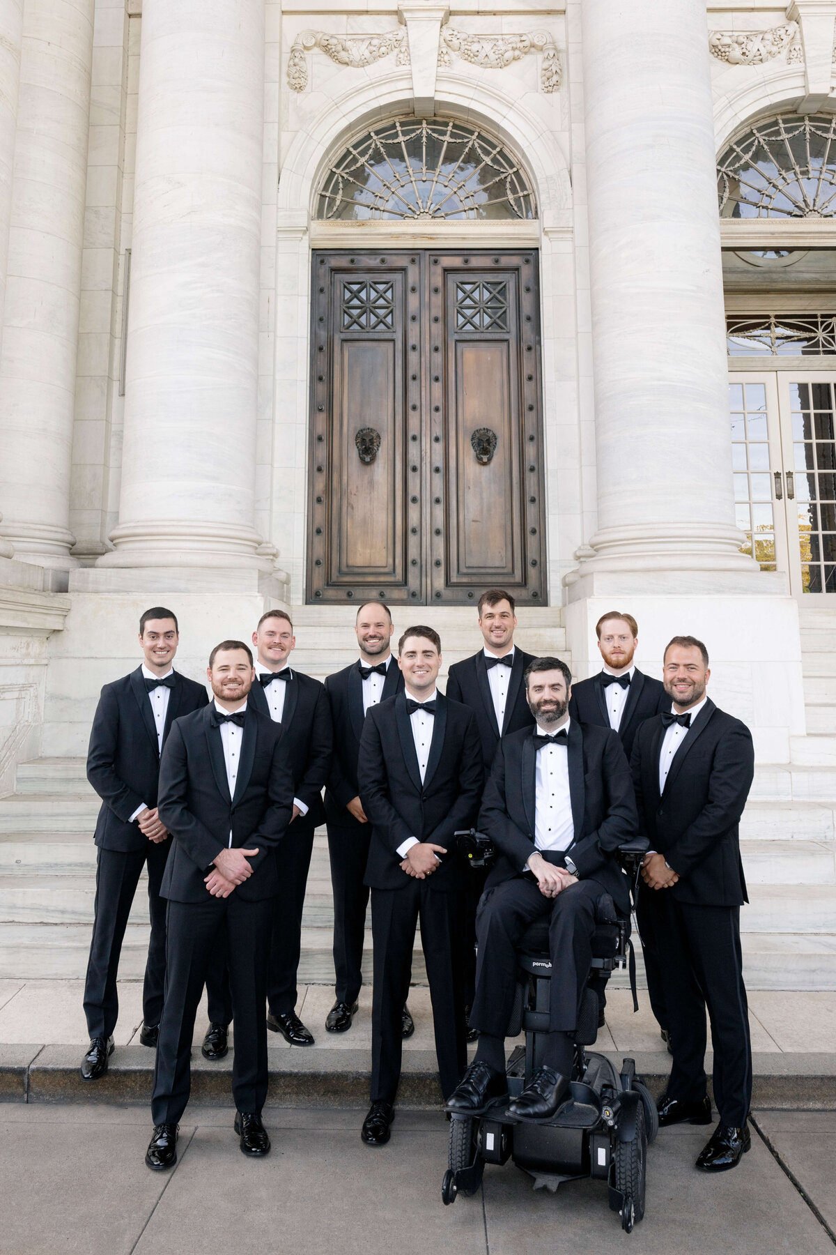 A group of nine men in tuxedos poses in front of a grand building with large double doors and columns. One man is seated in a wheelchair, and the others stand around him, all smiling.