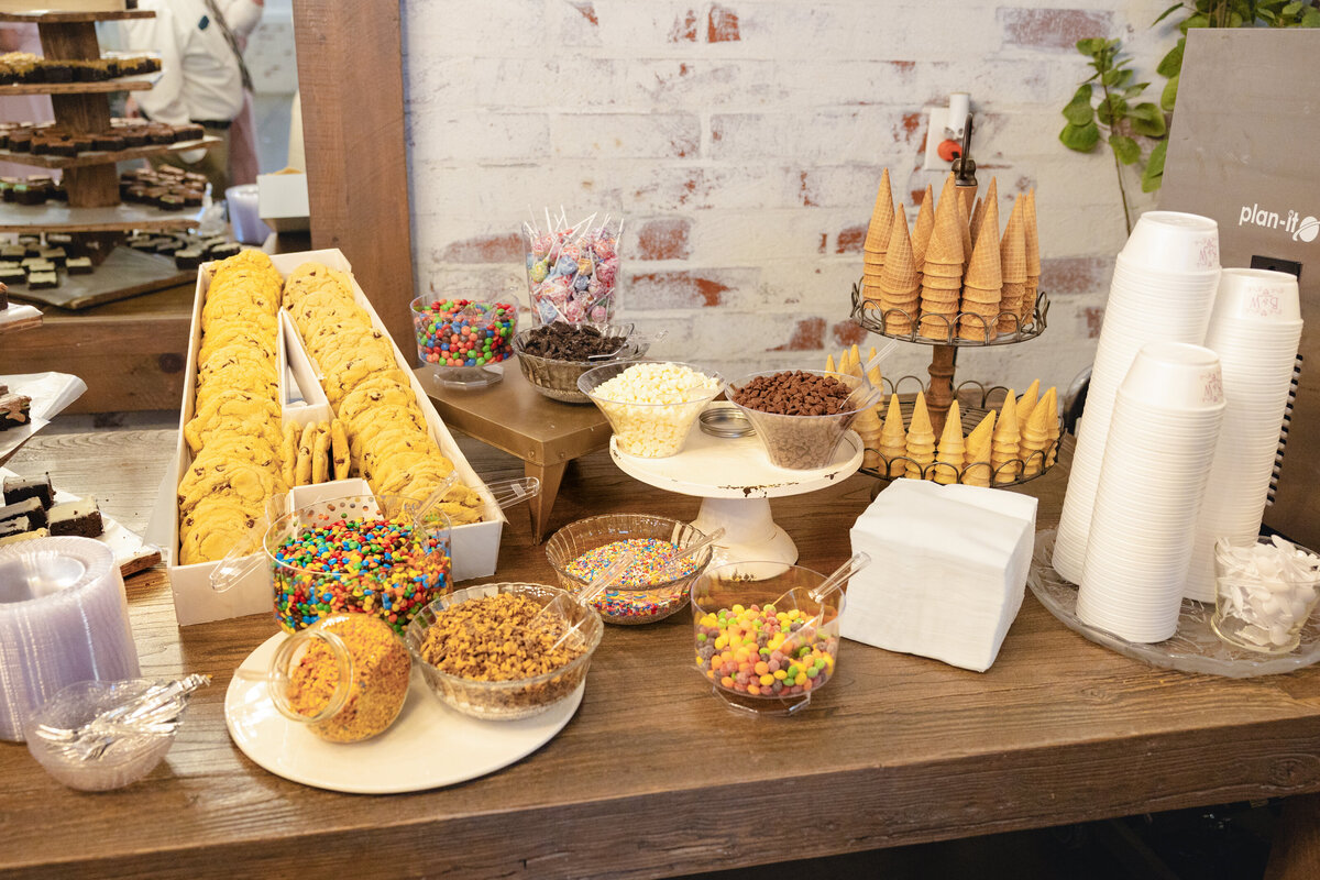 A beautifully arranged dessert bar featuring rows of cookies, waffle cones, and bowls of colorful ice cream toppings like sprinkles and chocolate chips.