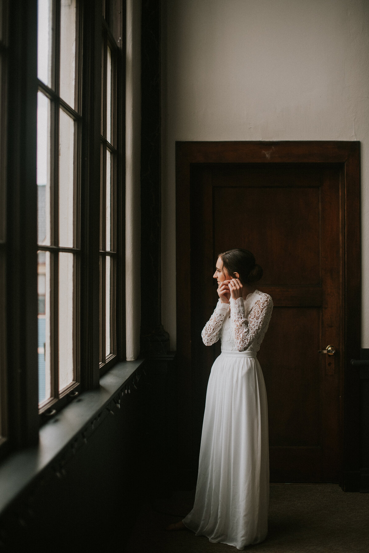 photo of a bride in a long sleeve dress getting ready putting her earrings on by a window at midtown center in holland michigan