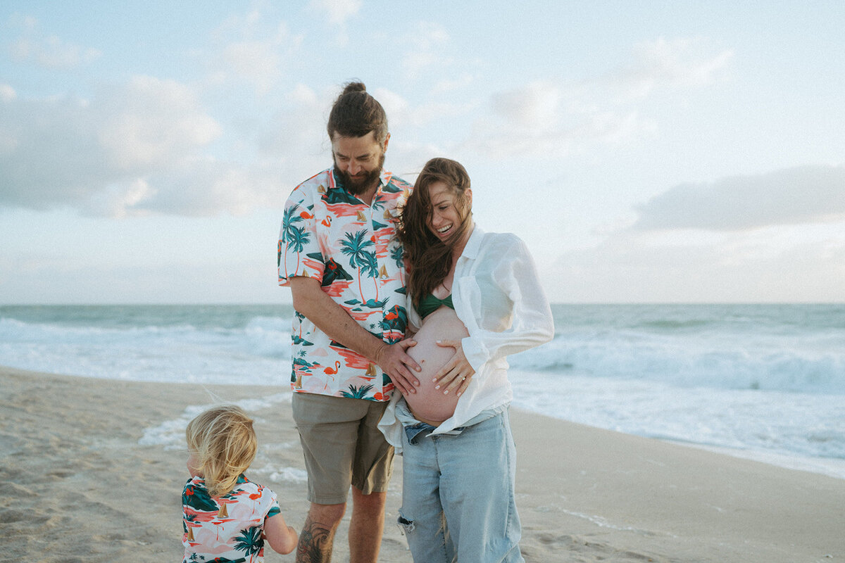 man touches woman's pregnant belly while son looks on during maternity photos at the beach captured by NYC maternity and family photographer Elsie Goodman