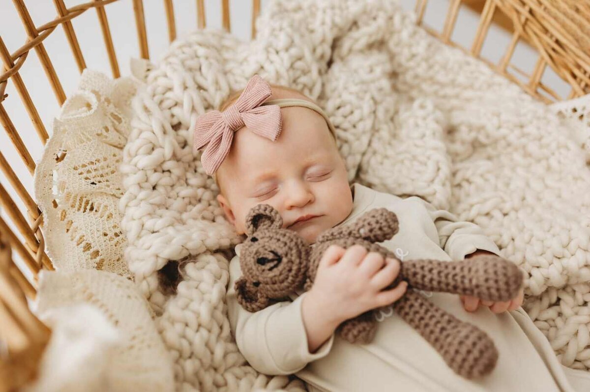 Ruua Hamdan Photography (UK): A baby with red hair sleeps peacefully, cuddling a small brown teddy bear, wrapped in a soft white blanket.