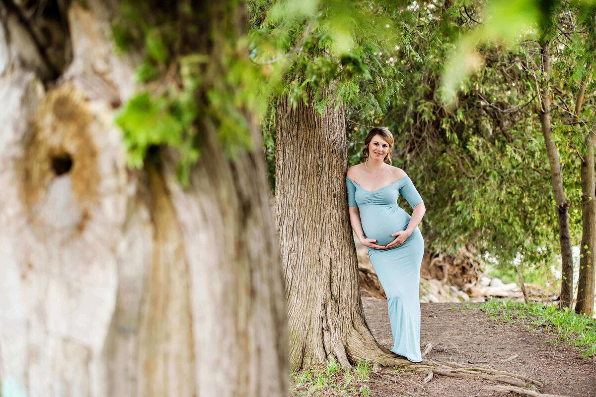 pregnant woman leaning against a tree wearing a blue maternity dress for her outdoor Burlington, Ontario maternity photography session