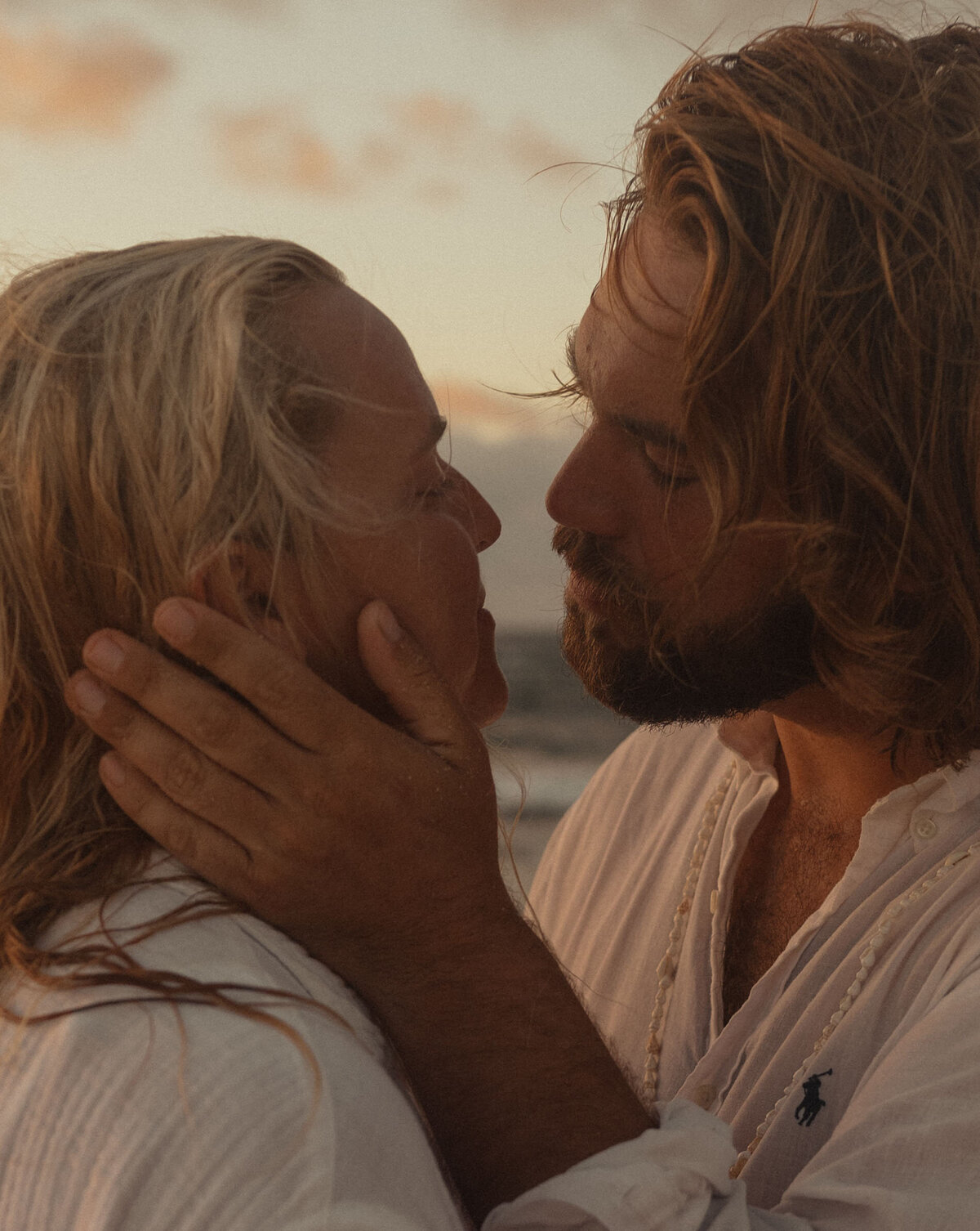 groom holds brides face in his hands as he goes in for a kiss while they stand in the ocean under a sunset sky