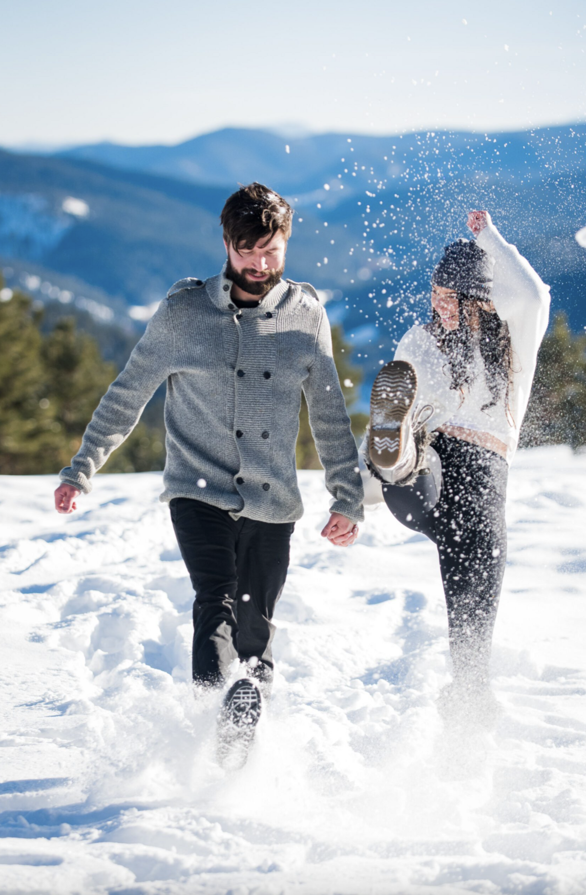 A man and woman playfully kick snow toward the camera during their engagement session at Mount Falcon in Colorado.