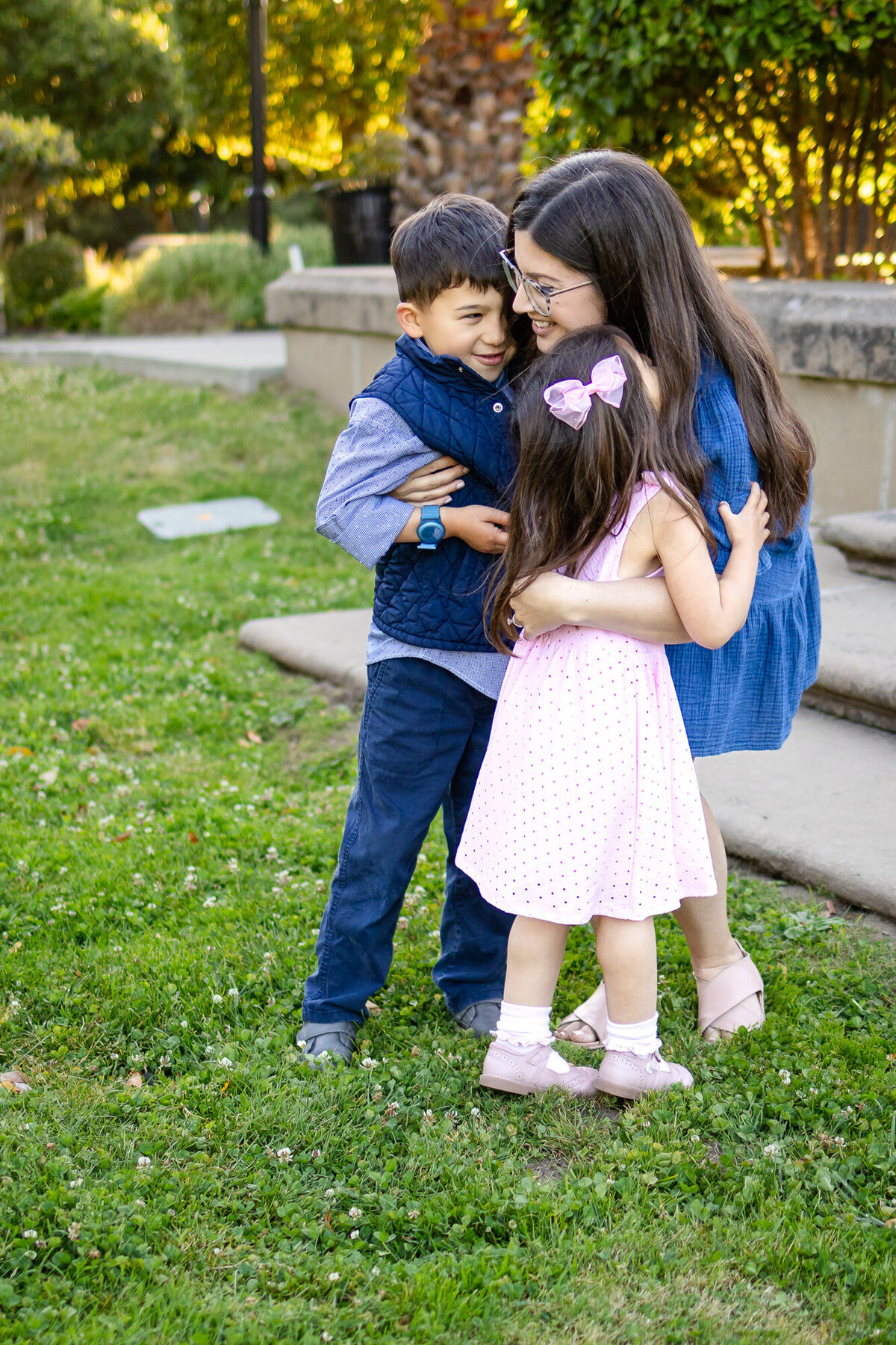 Bay Area Family Portfolio - Ellobelle Photography captures a heartwarming group hug between a mother and her two children in a grassy park with golden sunlight filtering through trees.