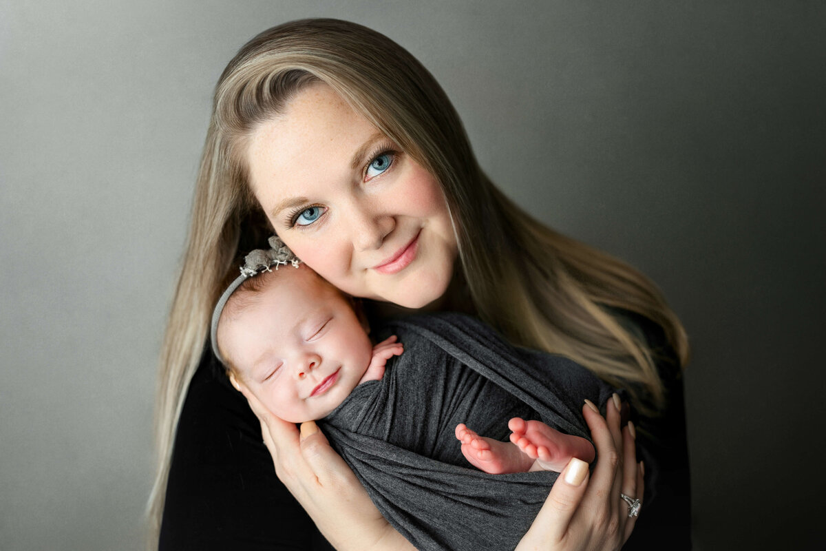 new mom holding her smiling baby girl on a grey backdrop  at a newborn photo shoot in Northern VA