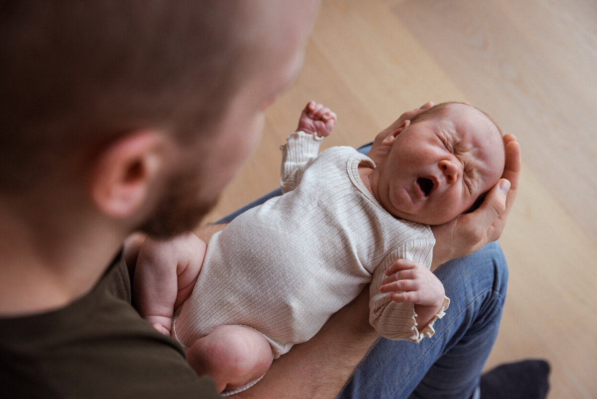 Livsstilsfoto av en baby som ligger på fars knær og gråter. Tatt ovenfra og ned.
