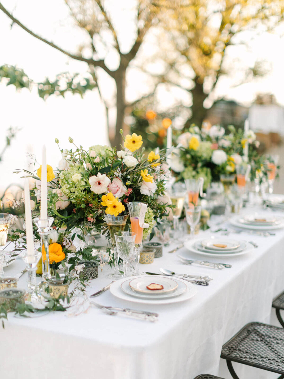 Elegant outdoor dining table adorned with lush yellow and white floral arrangements, candles, fine china, and crystal glassware.