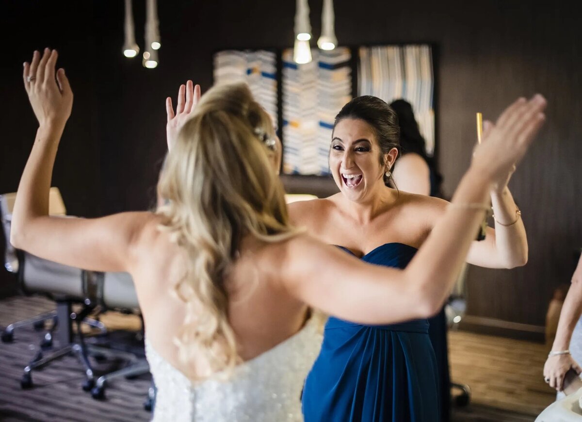 A candid moment before the wedding ceremony, where a woman in a blue dress raises her hands in excitement while facing the bride, symbolizing joyful anticipation.