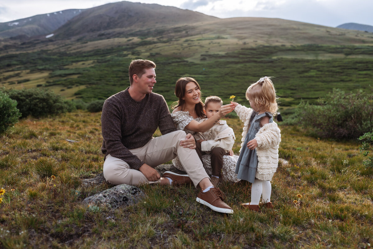 Little girl hands her mom a flower while the family of 4 snuggles in the mountains.