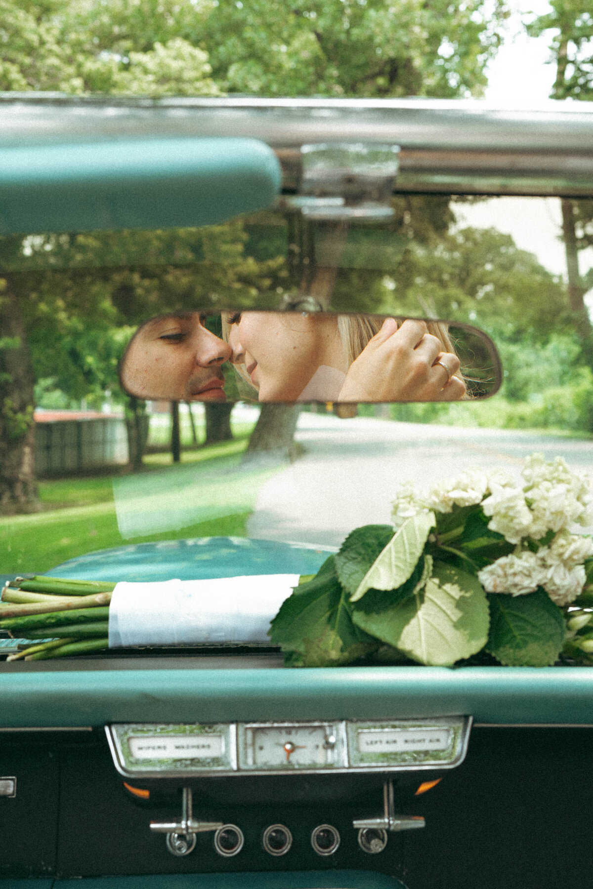 Bride and groom reflected in a vintage rearview mirror, sharing a quiet, romantic moment after their wedding drive—timeless, cinematic, and full of feeling.