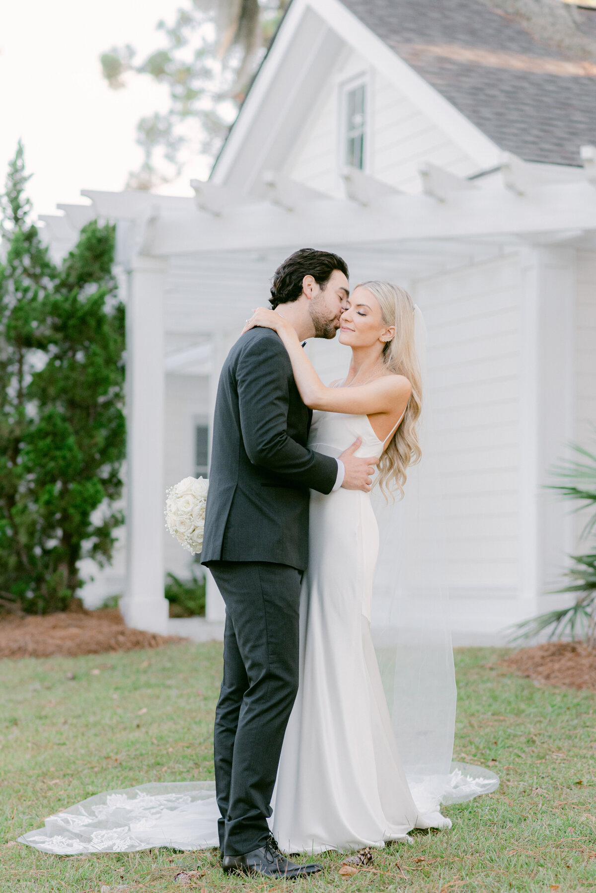 Groom kisses his bride, dressed in a sleek satin gown, outside a white pergola on Hilton Head Island—captured by luxury wedding photographer Amia Marcell.