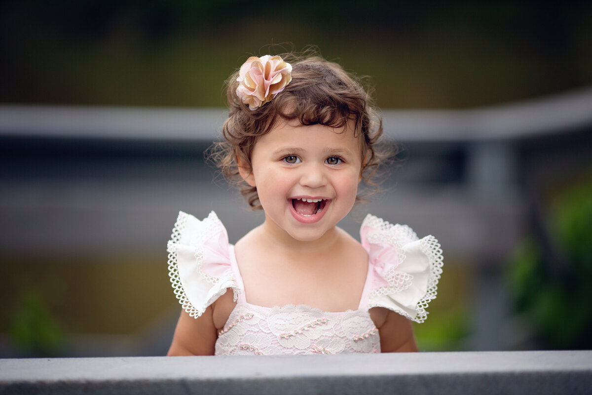 A happy young girl laughs while standing in a pink dress on a park bridge