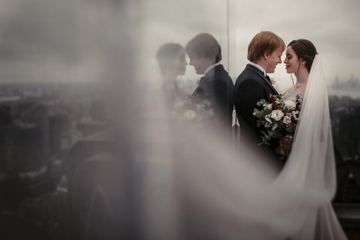 A groom leaning on the side of a glass building as his bride leans in for a kiss.