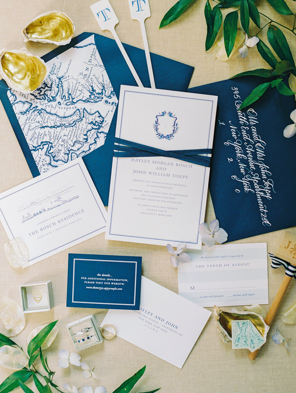 The photo was taken at The Hamptons, New York, waterfront estate tented wedding by photographer Bonnie Sen, capturing a stunning flat lay of an elegant wedding invitation suite. The invitations are navy blue and ivory, adorned with a monogram and ornate borders. Surrounding the invitations are detailed inserts, RSVP cards, decorative shells, lush greenery, and small accessories, evoking a sense of sophistication and style. The videographers for the event were East West Films.