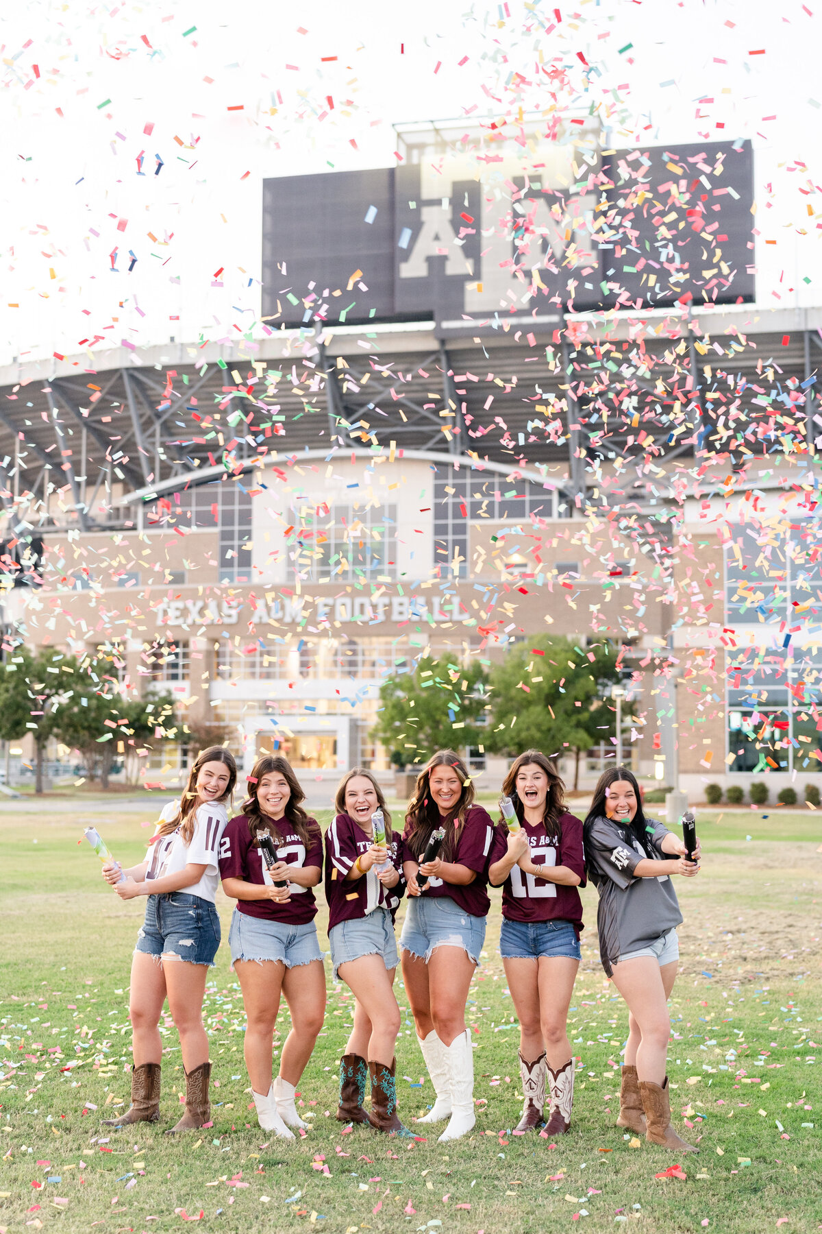 Texas A&M friend group wearing school jerseys, jean shorts and boots and popping confetti and laughing in front of Kyle Field